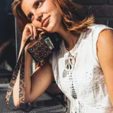 Llavero para el cuello con cinta de tela estampada con flores en tonos marrones, celestes y blancos. Tiene un mosquetón metálico negro y un aplique de cuero sintético negro con el logo de la marca grabado.