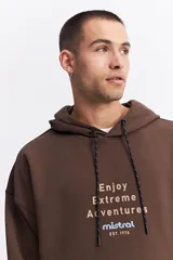 Canguro oversize color marrón, con capucha, cordones ajustables, bolsillo frontal con cierre y estampa frontal con texto y logo de la marca.