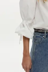 Camisa blanca de manga larga abullonada con detalle de punto smock en los puños y cordones para atar. Presenta cuello clásico y cierre frontal de botones.