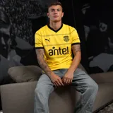 Camiseta de fútbol Puma del Club Atlético Peñarol, modelo Away 26, para hombre. Diseño amarillo con cuello en V y puños negros, y finas rayas horizontales negras en el frente. Incluye logos de Puma, Peñarol y Antel.