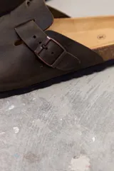 Zueco de cuero color marrón oscuro, con diseño de punta redonda, ajuste mediante hebilla metálica lateral y plantilla de corcho con suela de goma negra.