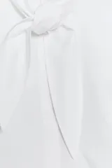 Camisa sin mangas confeccionada en algodón, con cuello redondo y detalle de lazada frontal. Presenta cierre frontal con botones ocultos por solapa y bajo redondeado.