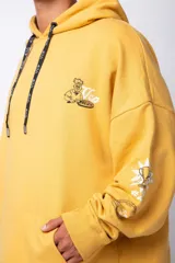Buzo canguro amarillo ocre oversize de algodón rústico con capucha, cordones negros con letras blancas, bolsillo frontal y puños y cintura acanalados. Presenta estampados en el frente y las mangas.