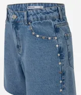 Short midi de jean azul con tiro medio, bolsillos frontales y traseros, trabillas para cinturón y detalle de tachas metálicas aplicadas en los laterales y bolsillos. El diseño se completa con un dobladillo deshilachado.