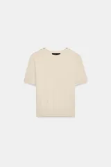 Jersey de punto de manga corta y cuello redondo, color beige, con detalle de botones dorados en los hombros.