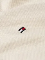 Chomba color beige de Tommy Hilfiger, modelo 1985, de corte regular y mangas cortas. Presenta cuello tipo chomba, cartera de dos botones, diseño de rayas en el interior del cuello y logo bordado en el pecho.