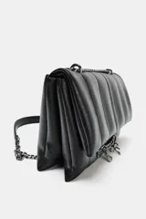 Bolso bandolera de diseño acolchado en color negro, con solapa frontal decorada con una cadena metálica y charms colgantes. Cuenta con cierre de botón imantado, triple compartimento interior y correa ajustable que permite llevarlo al hombro o cruzado.