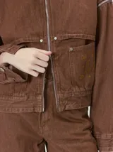 Campera de denim grueso color marrón, con cuello de pana y cierre frontal. Cuenta con mangas desmontables mediante cierre en los hombros, canesú aplicado en el pecho y dos bolsillos de parche cuadrados en el frente.