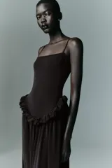 Vestido largo de color negro con escote recto y tirantes finos ajustables. Presenta un diseño con detalle de volados en la cintura que genera un efecto peplum y una falda con caída fluida.