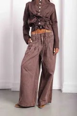 Pantalón de jean color marrón con silueta wide leg y tiro medio. Presenta una cintura trabajada con pasacintas entrelazadas, cierre frontal, bolsillos laterales y pinzas frontales que aportan volumen y caída a la pierna.
