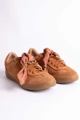 Zapatillas de gamuza color tostado con suela de goma a tono. Presentan un diseño con cordones tradicionales y un detalle decorativo de lazo de tela satinada con herrajes metálicos dorados en la parte superior.