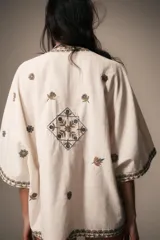 Blusa de escote en pico y manga corta, confeccionada en tejido color crudo con detalles de bordados florales y geométricos a contraste. Presenta cierre frontal con botones y bolsillos de plastrón en el delantero.