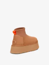 Bota corta tipo botineta de la marca UGG, color negro, con diseño de caña baja y plataforma elevada. Presenta una tira de agarre en color amarillo neón en la parte posterior y una suela de goma gruesa con textura acanalada.