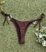 Conjunto de bikini color marrón chocolate, con corpiño triangular y bombacha colaless con tiras laterales para atar. Ambas piezas cuentan con argollas metálicas decorativas en el centro y los laterales.