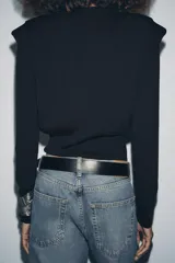 Jersey negro de punto, manga larga, con cuello redondo y detalle de pliegues o volados en la zona de los hombros.