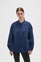 Camisa de jean azul con cuello mao, cierre frontal con botones y mangas largas con puños elásticos fruncidos.