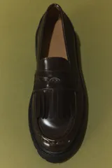 Mocasines tipo penny de color marrón oscuro, confeccionados en material sintético con acabado brillante. Presentan flecos decorativos en el empeine, una banda transversal con detalle metálico y una suela gruesa dentada de color negro.