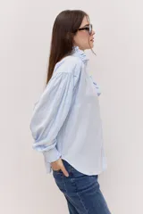 Camisa celeste con cuello alto, volados frontales y mangas largas con puños ajustados.