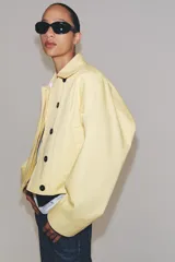 Chaqueta corta confeccionada en mezcla de algodón color amarillo claro, con cuello de solapa, manga larga, bolsillos delanteros y cierre frontal con botones visibles en los extremos.