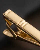 Clip de corbata metálico en tono dorado con diseño minimalista de líneas grabadas.