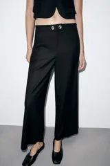 Pantalón negro de tiro medio con pierna ancha y cierre frontal de cremallera con dos botones dorados decorativos.