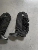 Botas estilo cowboy de color negro, con detalles de flecos largos en un lateral y bordados con apliques brillantes en la caña. Cuentan con un pie de cuero sintético y puntera fina.