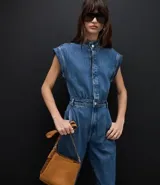 Mono utilitario de jean color azul medio, con cuello mao, cierre frontal oculto, diseño sin mangas con hombreras estructuradas, cintura entallada con trabillas y bolsillos funcionales.