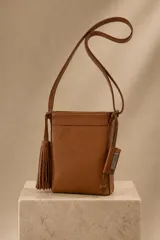 Bandolera de cuero color suela, de diseño rectangular vertical con cierre superior, correa larga ajustable y detalles de borla de flecos colgante en el lateral.