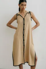 Vestido largo color beige con cuello camisero, sin mangas, abotonado al frente y con aberturas laterales. Presenta ribete negro en cuello, sisas, abotonadura y ruedo.