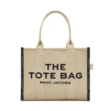 Bolso tote grande de lona color beige, con asas de mano y hombro. Presenta el texto "THE TOTE BAG MARC JACOBS" estampado en negro en el frente. Los laterales tienen tiras de tela negras con inscripciones blancas.