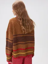 Pullover de punto con textura aterciopelada, diseño oversize, cuello polo con cierre frontal parcial y detalles de pespuntes en contraste. Presenta rayas horizontales en tonos tierra y un parche bordado con la letra M en el pecho.