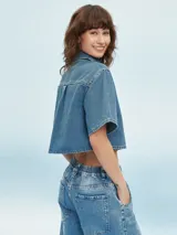 Camisa corta de denim azul claro, con cuello camisero, manga corta ancha y cierre frontal de botones blancos. Presenta un bolsillo de parche en el pecho izquierdo.