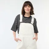 Enterito de jean color blanco con diseño de pierna ancha, bolsillo frontal en el pecho y tirantes ajustables con hebillas metálicas.