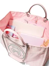 Bolso tipo shopper de Victoria's Secret, confeccionado en lona con diseño a rayas rosadas y blancas. Cuenta con asas de mano y correas de hombro con cadena metálica. Incluye un parche circular con el logo de la marca y alas bordadas en el centro.
