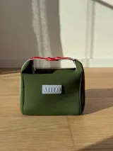 Neceser de neopreno color borgoña, con cierre superior y forro interior. Tiene un asa de mano con una franja azul y blanca.