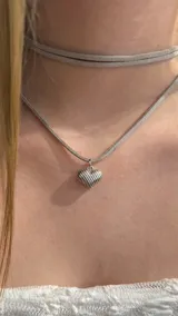 Gargantilla tipo choker de gamuza en color gris claro, con un dije metálico plateado en forma de corazón con diseño de líneas grabadas.
