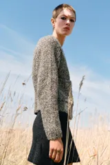 Chaqueta de punto de cuello redondo y manga larga, con textura jaspeada en tonos tierra y azul. Presenta cierre frontal con botones oscuros y dos bolsillos de plastrón en el delantero.