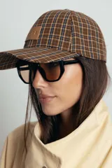 Gorra tipo baseball con estampado de cuadros escoceses en tonos azul, naranja y beige. Presenta un parche de cuero sintético en el frente y cierre ajustable en la parte posterior.