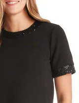 Blusa negra de manga corta con cuello redondo y puños decorados con ribetes de lentejuelas brillantes. Confeccionada en tejido suave y fluido, presenta una silueta recta y cierre de ojal con botón en la espalda.