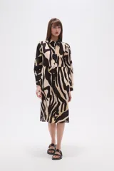 Vestido tipo camisa de corte holgado, con estampado de hojas en tonos marrones y beige. Presenta cuello camisero, cierre frontal con botones y mangas largas.