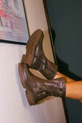 Bota de caña media confeccionada en cuero color marrón, con suela gruesa tipo tractor antideslizante. Presenta estribos decorativos con hebillas metálicas en el tobillo y una correa ajustable en la parte superior de la caña. Incluye cierre lateral para facilitar el calce.