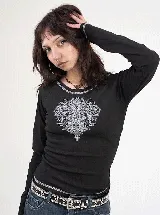 Remera de manga larga color negro, con corte ajustado y estampado frontal en serigrafía con diseño ornamental gótico. Presenta costuras decorativas visibles en el cuello y el ruedo.
