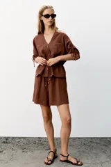 Bermuda marrón de lino, de corte holgado, con cintura elástica fruncida y cordón ajustable con abalorios en los extremos. Cuenta con bolsillos delanteros.