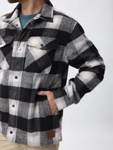 Sobrecamisa de estilo leñador con estampado de cuadros en tonos gris, negro y blanco. Presenta cierre frontal con cremallera y solapa con botones a presión, cuello clásico y bolsillos laterales.