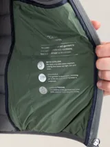 Chaleco acolchado de hombre color gris, con diseño matelasseado horizontal, cuello alto y cierre frontal completo. Presenta un calce ajustado y detalle de logo circular bordado en el pecho.