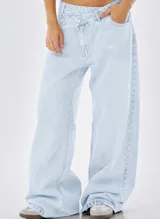 Jean baggy de denim celeste claro, con tiro medio y diseño de cintura cruzada asimétrica con cierre de botón único y bolsillos laterales.