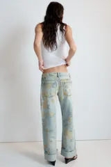 Jeans de tiro bajo confeccionados en denim, con diseño holgado y estampado integral de rosas en tonos amarillos y verdes sobre fondo celeste claro. Cuenta con cinco bolsillos, trabillas para cinto y botamangas con vuelta.