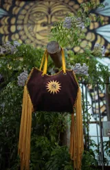 Bolso de gamuza color marrón oscuro con forma trapezoidal, asas de cuero amarillo mostaza y largos flecos amarillos en los laterales. Presenta un logo bordado en el centro que simula un sol con las iniciales "MS" o "1S1" en su interior.