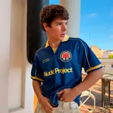 Camiseta de fútbol Nude Project color azul con cuello polo amarillo. Presenta detalles en amarillo en los hombros y mangas, estampado frontal con el nombre de la marca y un escudo circular con una rosa en el pecho.