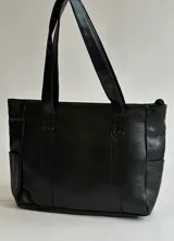 Cartera tipo shopper de cuero sintético negro, con doble asa larga para hombro, bolsillos laterales externos y cierre superior.